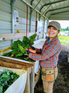 Reiden, a farmer enrolled in the Headwaters Farm Incubator program, washes vegetables while turning toward the camera and smiling. In the background a wall and roof overhang is visible, and the farm past that