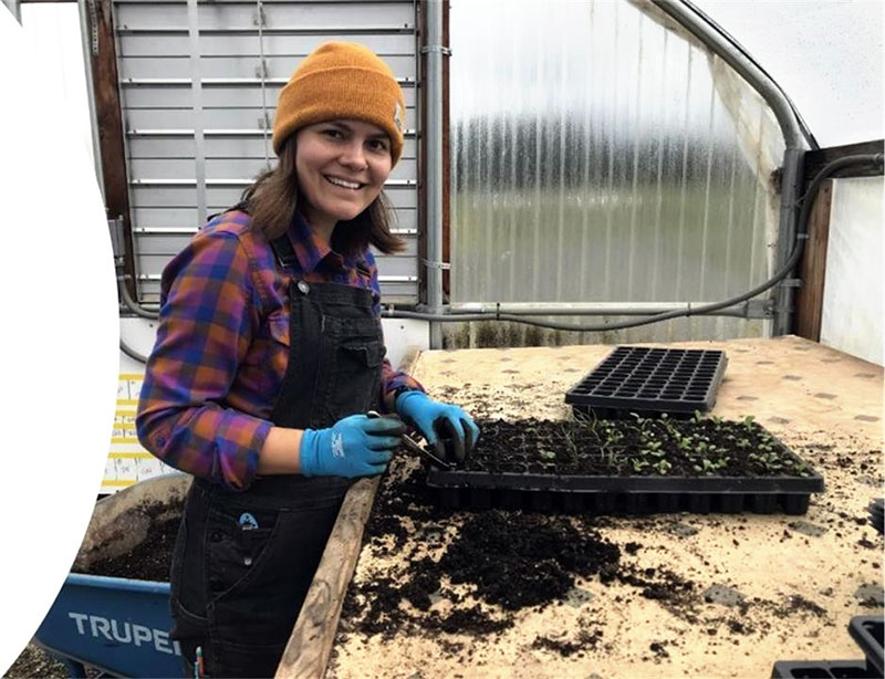 A Headwaters farmer wearing overalls and a beanie, with a plant propogation tray full of small plant starts in front of her on a wooden table. She turned toward the camera and smiling