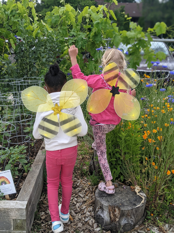 등에 장식용 꿀벌 날개를 달고 있는 두 명의 어린 아이들이 카메라를 바라보고 있는 초목과 정원 침대를 확인하고 있습니다