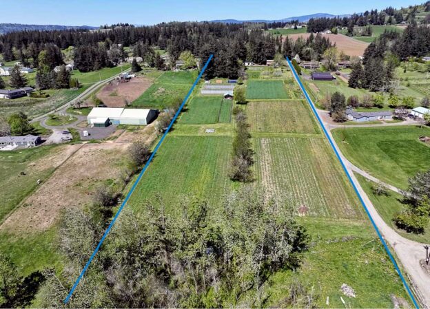 Foto aérea de la propiedad agrícola de Woodard Road, que muestra parcelas agrícolas, estructuras agrícolas, caminos y colinas boscosas al fondo. La propiedad en sí está marcada por dos líneas azules en el centro de la foto.