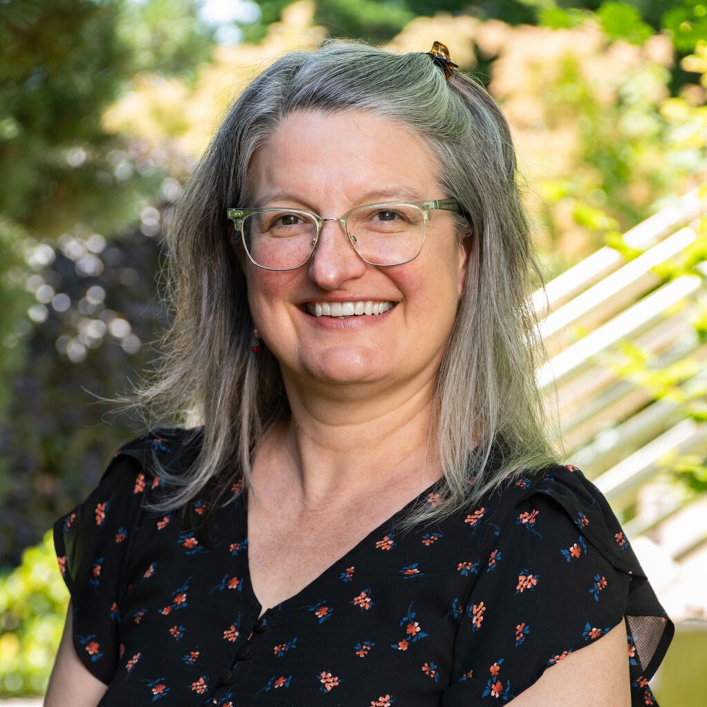 close-cropped profile portrait of Nikola, smiling in the foreground with stairs and foliage in soft focus in the background