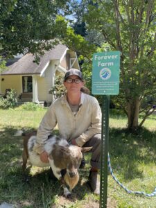 A person wearing glasses with hair pulled back under a ball cap kneels with a goat in front of a sign that says "Forever Farm"
