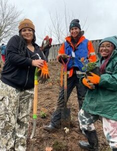 Three people pose for a photo while planting trees and shrubs. Two are holding shovels, and one is holding a plan.