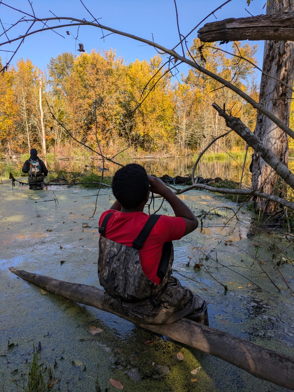 A young person in a red tee shirt sits on a log looking into the distance through binoculars.