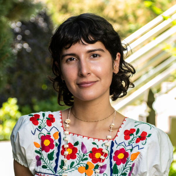 profile photo of Isa smiling, photo cropped to the shoulders, staircase and foliage visible in soft focus in the background
